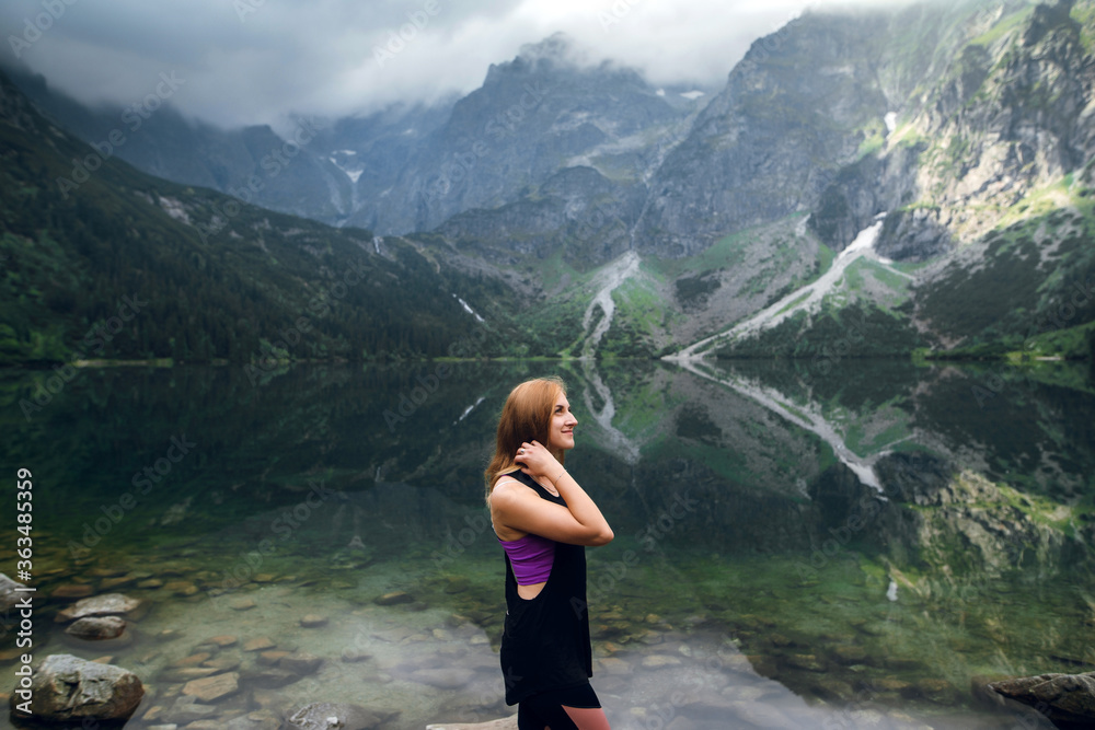 Beautiful young fit woman in sportswear standing on the rocky shore and admiring scenic view of green hills and mountains on Morskie Oko lake, High Tatras, Zakopane, Poland.