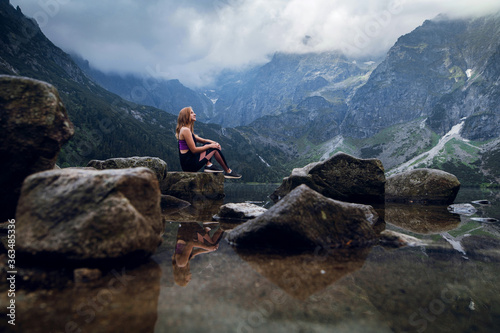 Young blonde fit and sporty woman in sportswear sitting on the rock on the shore and admiring beautiful view of green hills and mountains on Morskie Oko lake, High Tatras, Zakopane, Poland.