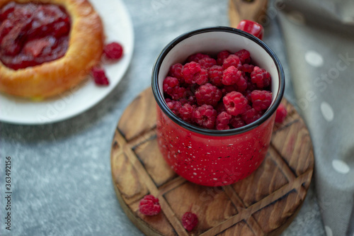 In the foreground is a red mug with garden raspberries, in the background a cheesecake with berry filling, light background, defocus; summer recipes, homemade vitamin snack