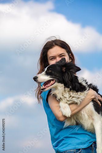 A girl with long hair in a blue T-shirt holds a black and white border collie dog in her hands and hugs. Portrait against a bright blue sky with clouds. Vertical orientation. 