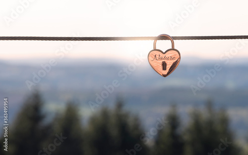 Copper-coloured lock in the shape of a heart with the inscription "Nature" hanging from a wire rope in front of a wooded landscape as a symbol for nature and environmental protection 