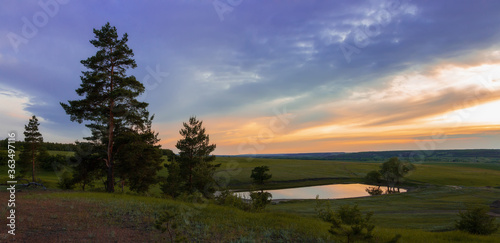 Lake at sunset in the forest among of green meadow and dramatic clouds. Picturesque summer landscape of Russian nature.