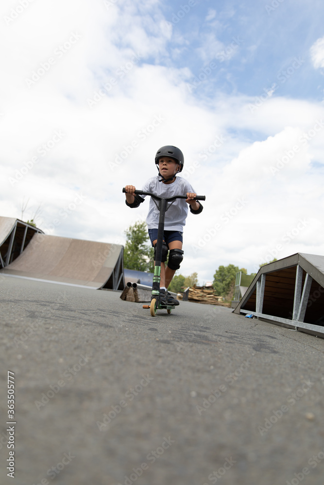 A cute little boy rides a scooter in a skatepark.