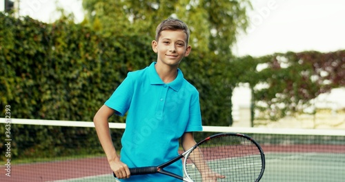 Portrait of teen small Caucasian cheerful boy in blue polo shirt holding racket and smiling happily to camera at sport court outdoor. Happy child spending day at sporty activity. © VAKSMANV