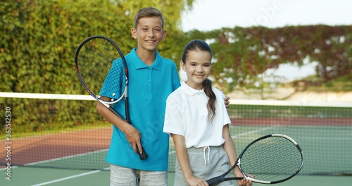 Portrait of Caucasian cute cheerful small kids holding rackets and standing at sport court on summer sunny day. Little joyfull tennis players smiling to camera. Friendly boy and girl at pitch.