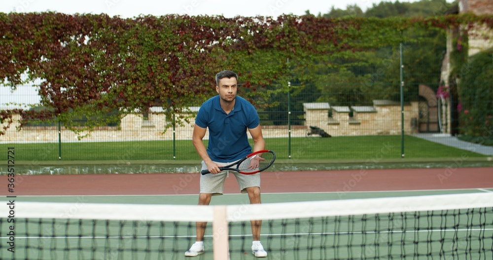 Caucasian cheerful handsome man playing tennis and hitting ball with racket on sunny summer day at sport court. Attractive male having fun at sporty game outdoors. Caucasian cheerful handsome man playing tennis and hitting ball with racket on sunny summer day at sport court. Attractive male having fun at sporty game outdoors.