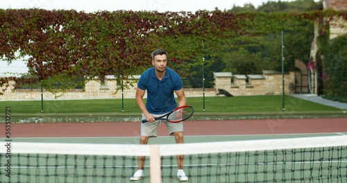 Caucasian cheerful handsome man playing tennis and hitting ball with racket on sunny summer day at sport court. Attractive male having fun at sporty game outdoors.