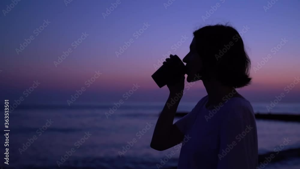 Silhouette of a girl at dusk drinks enjoying coffee on the sea at the beach. Atmospheric VioletSky Slow motion Silhouette of a girl at dusk drinks enjoying coffee on the sea at the beach. Atmospheric VioletSky Slow motion
