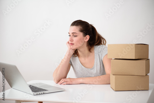 The girl sits thoughtfully at a laptop while supporting her chin with her fist. Near a stack of cardboard boxes on the table © maxfotoadobe