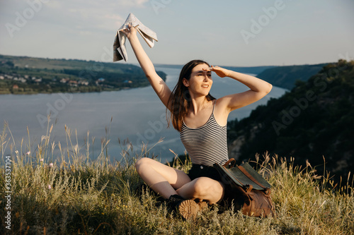 Young woman sitting on a cliff with a view, holding a map, waving hello to her friends.