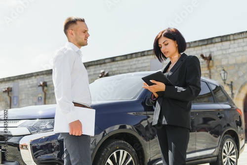 Partners, greeting. Young adult smiling man and woman in business dark suits standing near a car, shaking hands © Serhii