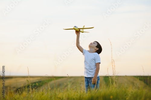 Happy child playing with a toy plane in nature during summer sunset. Boy in a white shirt with a plane in hands on wheat field. Kid holds a wooden airplane and dreams of being a pilot, on the nature © Serhii