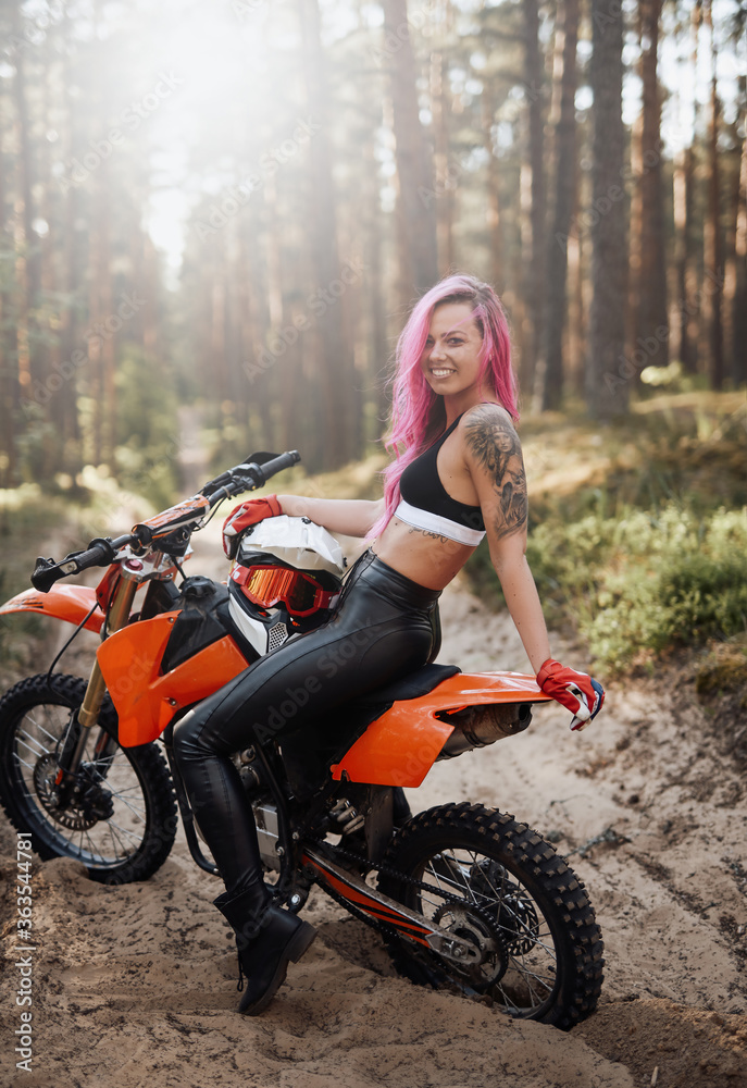 Beautiful young hipster girl with pink hair and tattoo on hand sitting on her modern motocross bike in woods, smiling and looking on the camera, illuminated by bright sunlight Beautiful young hipster girl with pink hair and tattoo on hand sitting on her modern motocross bike in woods, smiling and looking on the camera, illuminated by bright sunlight