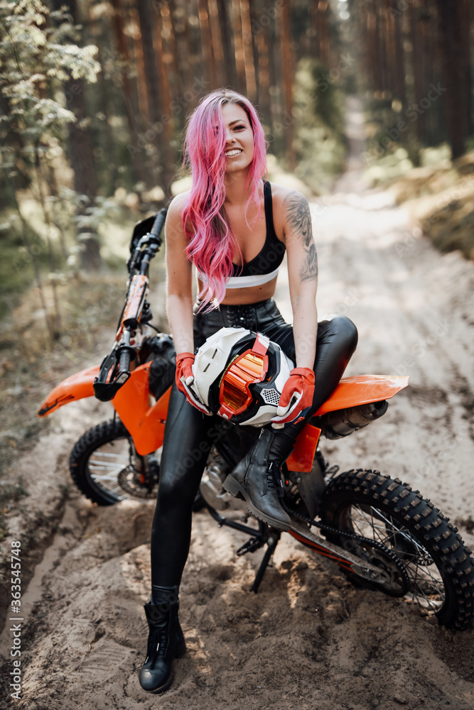 Happy young woman with pink hair and tattoo on hand sitting on her modern motocross bike in woods