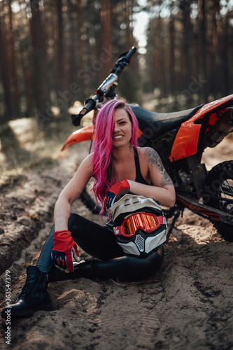Portrait of a charming young female racer with pink hair and tattoo on hand, smiling and looking on the camera while sitting on sand next to motorcycle in off road adventure