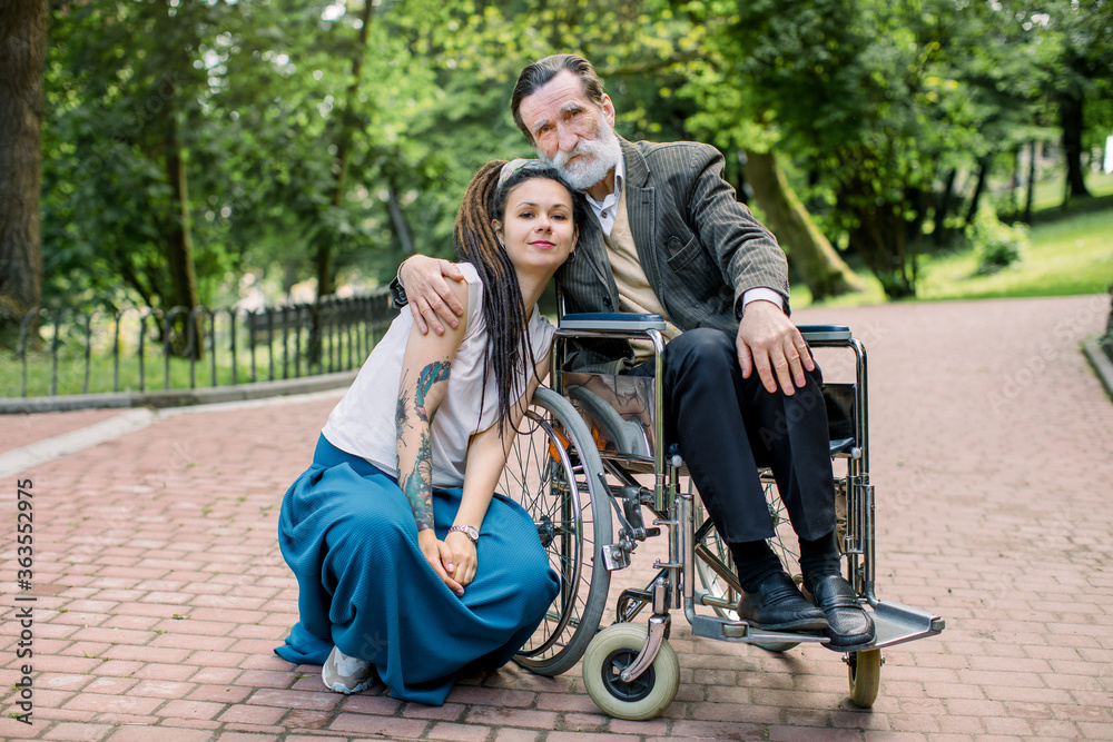 Obraz premium Young social worker or granddaughter takes care about senior man in wheelchair at walk in a park, smiling and embracing each other, looking at camera. Different generations of people