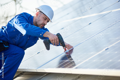 Male engineer in blue suit and protective helmet installing solar photovoltaic panel system using screwdriver. Electrician mounting solar module on roof of house. Alternative energy ecological concept