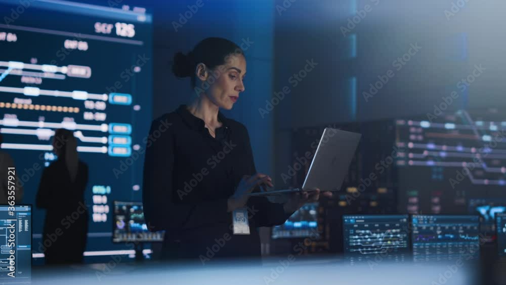 Beautiful Confident Female Project Manager works on Laptop Standing in Telecommunications System Control Room. In Background Big Screen Showing Infographics of Infrastructure, Charts, System Analysis