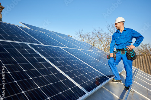 Happy man worker in blue suit and protective helmet installing solar photovoltaic panel system. Professional electrician standing on roof of house. Alternative energy ecological concept. Copy space