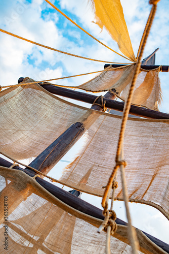 Bottom view of a ship mast with beige sails swings against a blue sky with sunny sunny summer warm day clouds. Marine and adventure concept