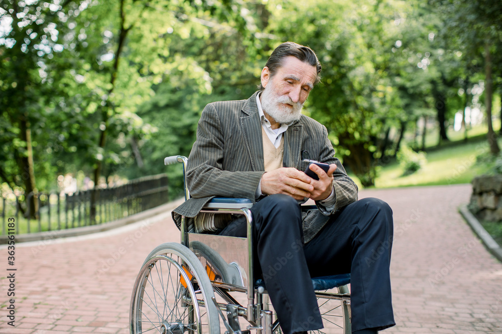 Bearded Caucasian old man is sitting in a wheelchair in the park alone, using his smartphone for serfing internet or socail networks Bearded Caucasian old man is sitting in a wheelchair in the park alone, using his smartphone for serfing internet or socail networks