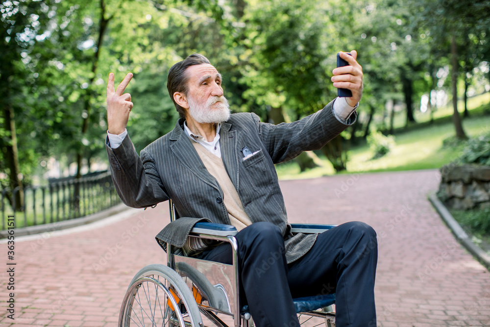Happy senior disabled or handicapped bearded man sitting on a wheelchair in park, using his smartphone for photographing himself on the background of park landscape Happy senior disabled or handicapped bearded man sitting on a wheelchair in park, using his smartphone for photographing himself on the background of park landscape