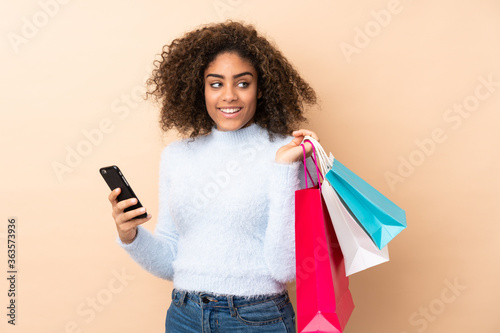 Young African American woman isolated on beige background holding shopping bags and a mobile phone