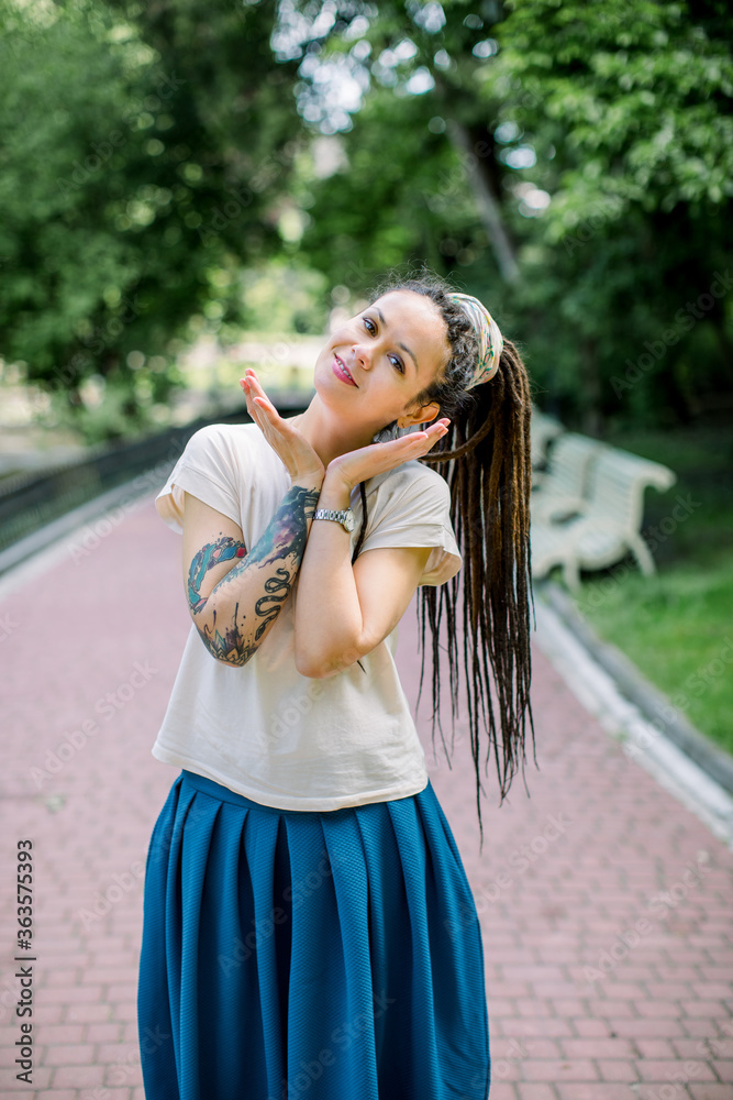 Naklejka premium Creative hipster girl with dreadlocks and tattoos in a city park on sunny summer day. Young stylish girl with dreadlocks outdoors