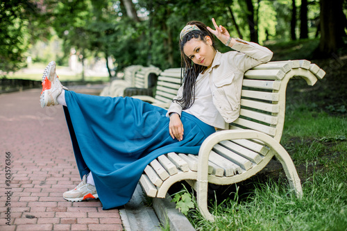Attractive young hipster woman with dreadlocks sitting on the bench in city park and ooking at camera, keeping her leg raised and showing victory sign. Freedom concept