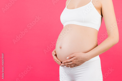Cropped image of pregnant woman in white underwear embracing her big abdomen at pink background. Maternity concept. Copy space © sosiukin