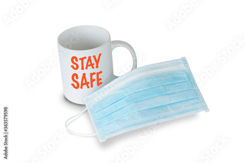 A light blue medical face mask in front of a white mug with red text saying STAY SAFE on white background. Prevention of Coronavirus Covid-19 concept.