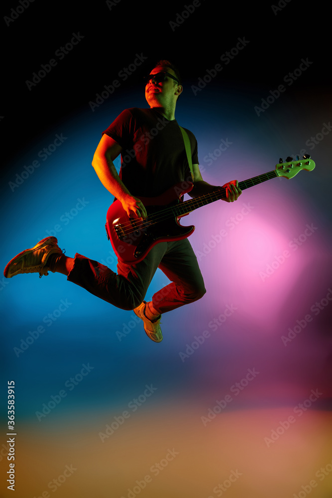 Jumping high. Young inspired and expressive musician, guitarist performing on gradient colored background in neon. Concept of music, hobby, festival, art. Joyful artist, colorful, bright portrait.