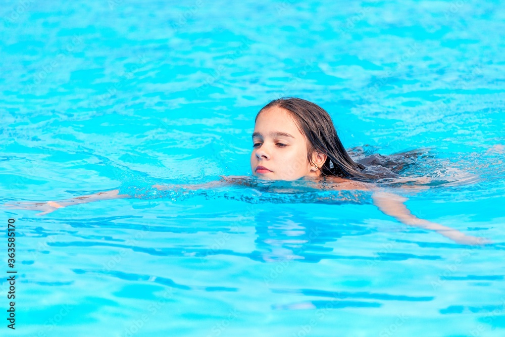 Teenage girl swims in the clear blue water of a pool during a vacation in a warm tropical country on a sunny warm summer day. Travel concept. Advertising space