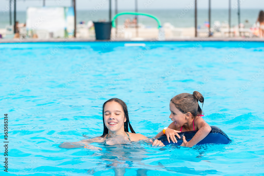 Fototapeta premium Two little sisters girls are swimming in a large pool with clear blue water of mine near the hotel on the background of the sea and the beach. Concept vacation tropical hot country with children