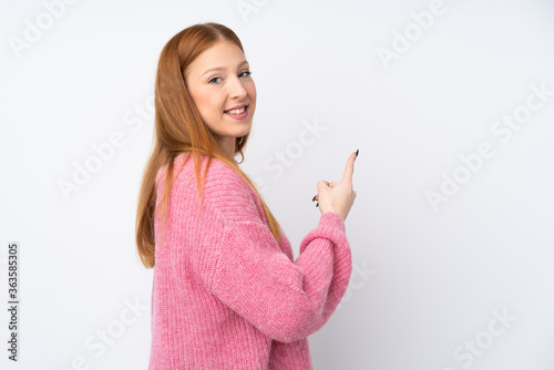 Young redhead woman with pink sweater over isolated white background pointing back