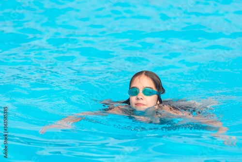 Charming girl teenager wearing waterproof glasses for the pool swims in the clear warm and blue water during the summer holidays. Concept of physical activity for children. Advertising space © YouraPechkin