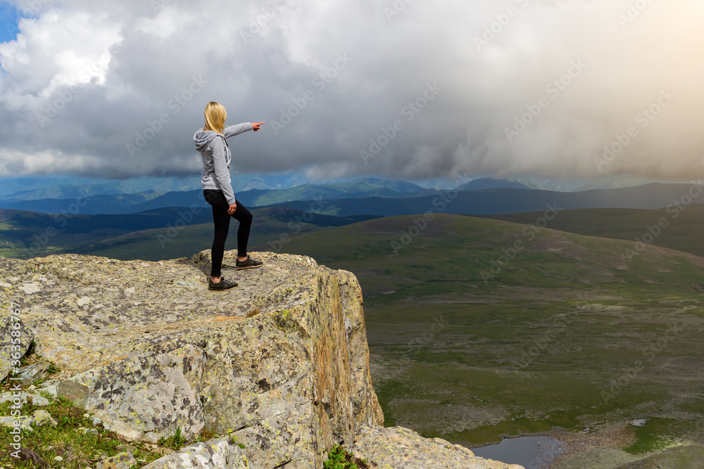 Young blonde girl in gray sweatshirt with black leggings stands on a cliff in the Altai mountains and points outstretched finger to the horizon where there is a picturesque landscape with many hills.