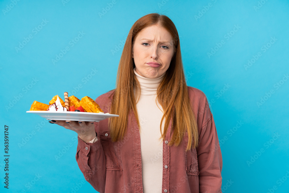 Young redhead woman holding waffles over isolated background with sad expression Young redhead woman holding waffles over isolated background with sad expression
