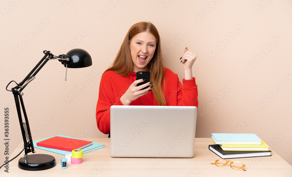 Young student woman in a workplace with a laptop with phone in victory position Young student woman in a workplace with a laptop with phone in victory position