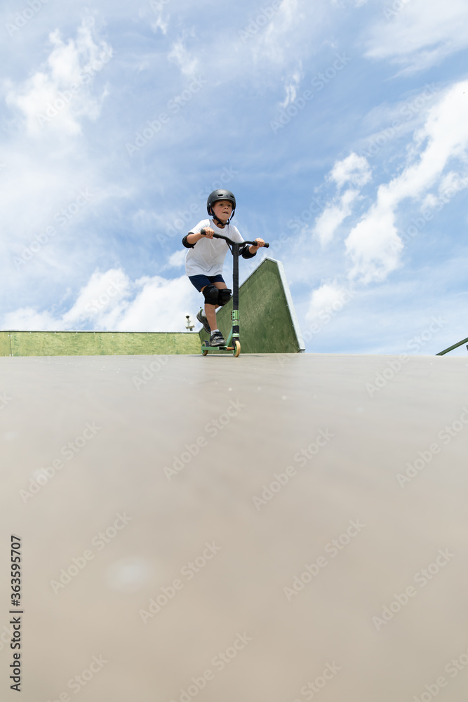 A cute little boy rides a scooter in a skatepark.