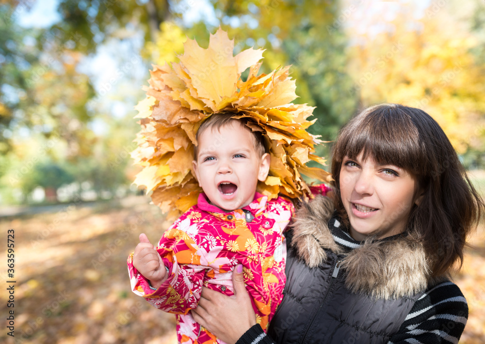 Fototapeta premium Charming young mother holds in her arms a pretty little girl with a wreath of yellow maple autumn leaves on her head while walking in the park. Autumn concept