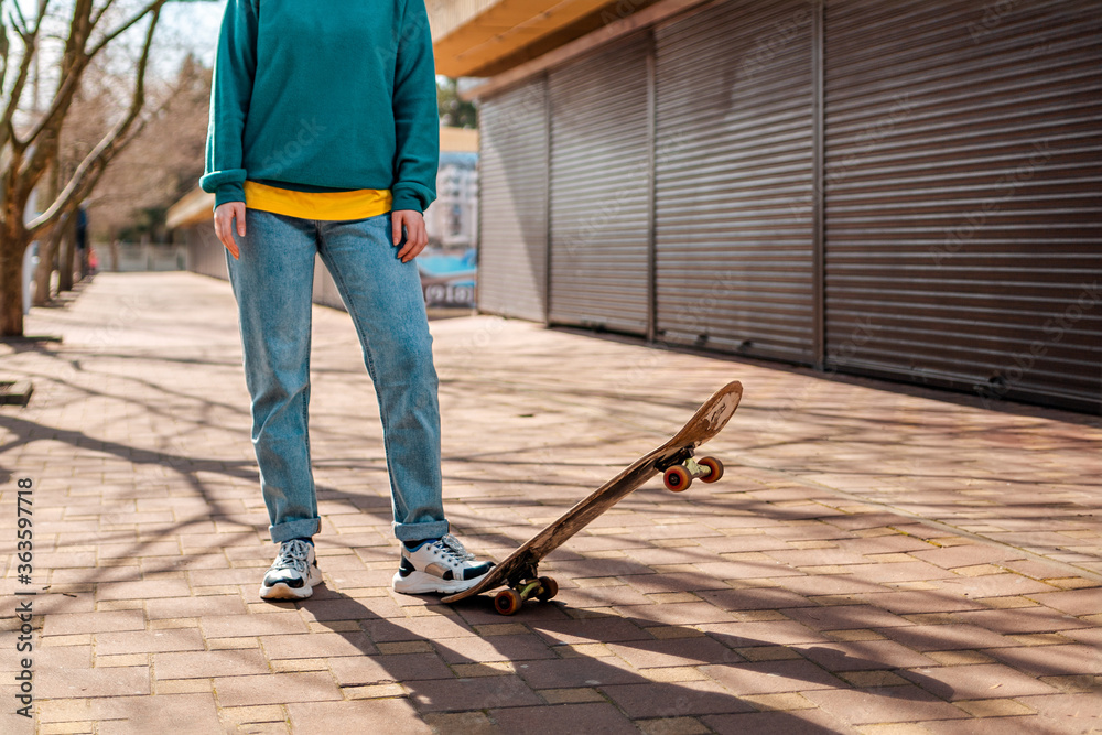 A hipster teen poses with her foot on a skateboard and looks into the distance. In the background, an alley. Close up. Concept of sports lifestyle and street culture A hipster teen poses with her foot on a skateboard and looks into the distance. In the background, an alley. Close up. Concept of sports lifestyle and street culture
