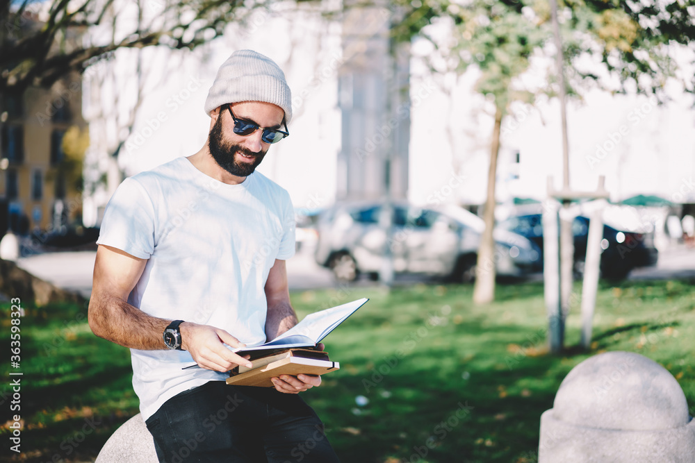 Positive handsome male student reading book enjoying learning outdoors on college campus, young bearded hipster guy spending time on hobby sitting near copy space area for your advertising content