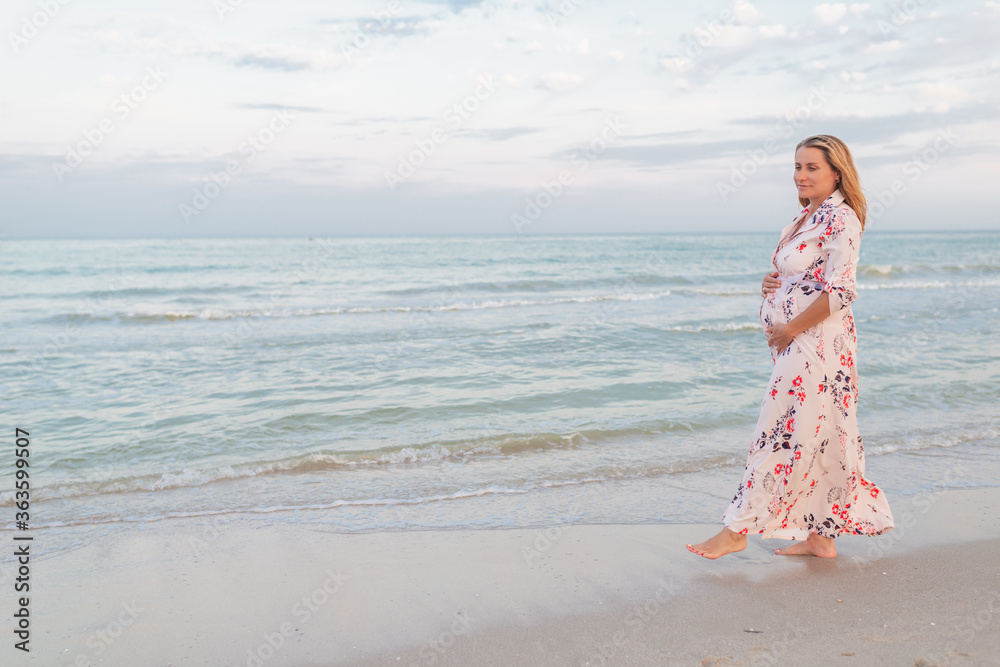 Beautiful happy young pregnant woman in a summer light dress hugging her belly and walks along the sea waves on a sunny summer evening during vacation. Natural Parenthood Concept. Copyspace Beautiful happy young pregnant woman in a summer light dress hugging her belly and walks along the sea waves on a sunny summer evening during vacation. Natural Parenthood Concept. Copyspace
