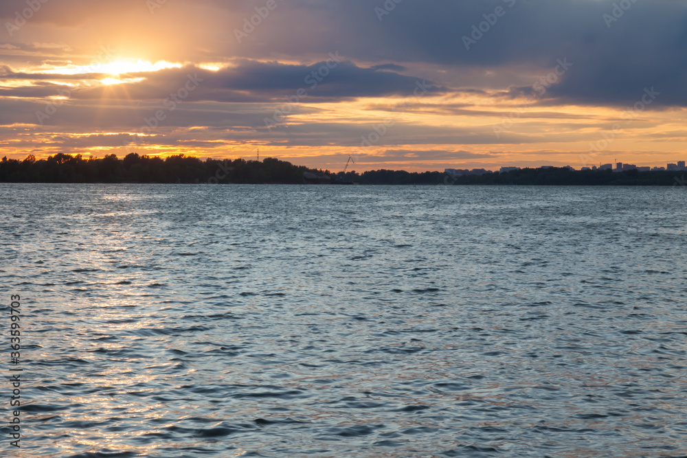 Evening landscape with the setting sun on a background of a painted dark sky in blue and orange colors in the clouds above a river or lake with reflecting rays in the water in the countryside.
