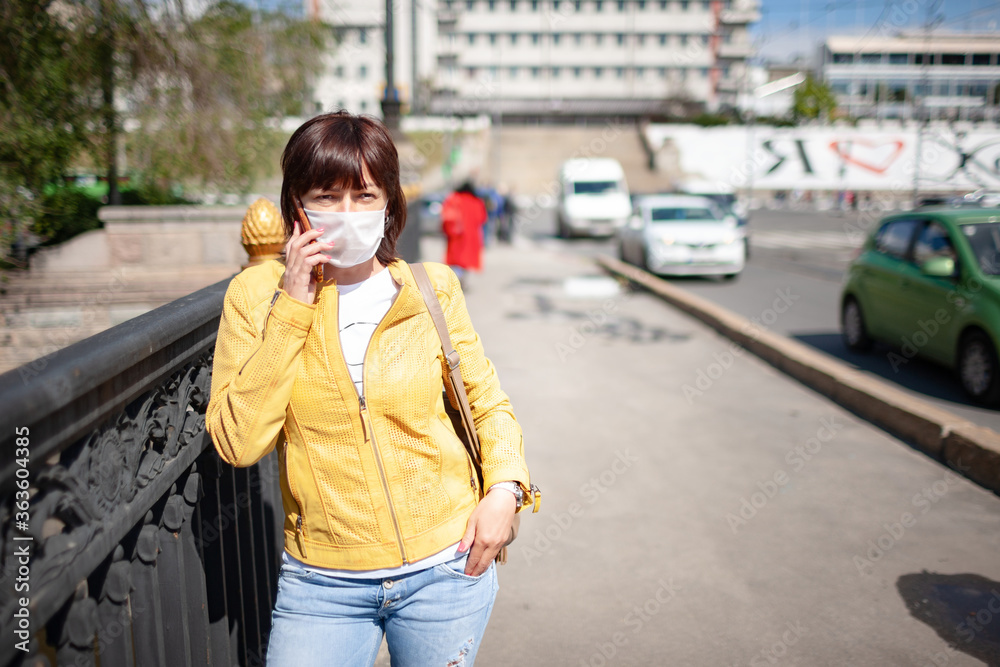 Funny middle-aged woman in a white protective mask talking on a smartphone while walking around the city on a warm spring day. Coronovirus pandemic and remote communication concept