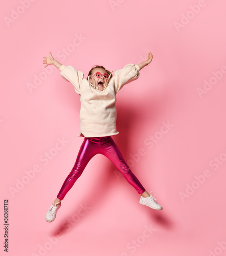 Fotografi Cute little girl in stylish casual attire jumping up spreading arms and legs widely and shouting in delight