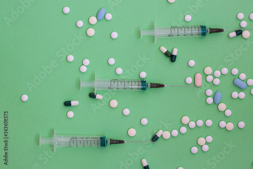 Three syringes with a needle empty lying on a green background and a lot of pills around