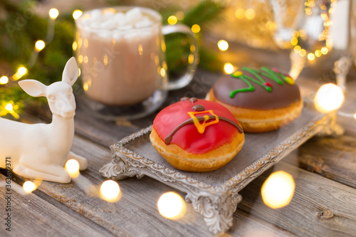 Christmas chocolate donuts with Santa Claus icing and new year tree on ceramic stand. Glass cup of cocoa with marshmallows on wooden table. Spruce branch, present, cone, sugar bowl,white deer,garland