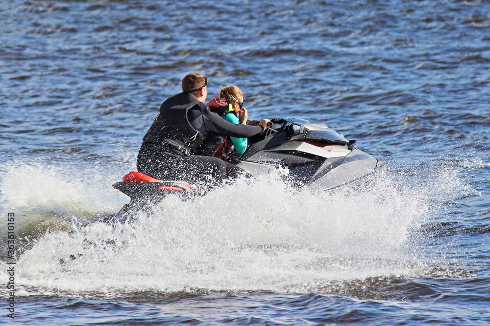 Modern powerful aquabike boat fast floating on blue water with many splash on summer day, jetski outdoor active sports recreation together man and girl couple people pair in black lifejackets Modern powerful aquabike boat fast floating on blue water with many splash on summer day, jetski outdoor active sports recreation together man and girl couple people pair in black lifejackets